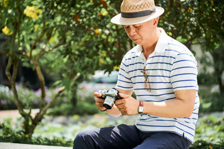 Senior man in hat sitting on bench in the park and looking just made photos on his photo cameraの写真素材