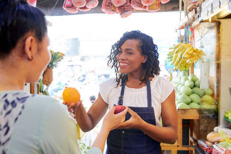 Smiling pretty female fruit vendor helping customer with choosing best fruitsの写真素材