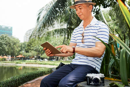 Asian senior photographer sitting with photo camera on the bench and working with photos online on digital tablet in the park outdoorsの写真素材