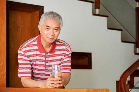 Portrait of Asian mature man with grey hair and in casual shirt standing with glass of water and looking at cameraの写真素材