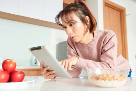 Asian young girl using digital tablet for online work while eating cornflakes with milk and fresh apples during breakfast in the kitchenの写真素材