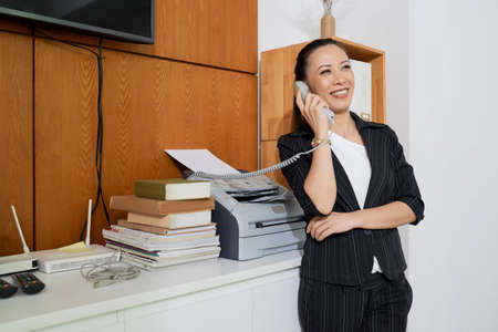 Asian young businesswoman standing in formal wear consulting somebody on the phone and smiling while working at officeの写真素材