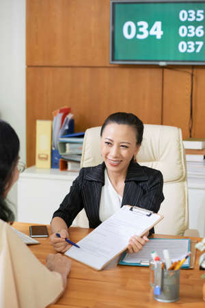 Asian young social worker smiling to her client and pointing at business contract and suggesting her to conclude a dealの写真素材