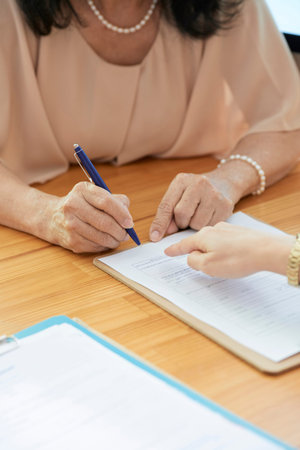 Close-up of mature businesswoman sitting at the table and signing a business contract with pen with...