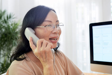 Mature businesswoman in eyeglasses holding handset and discussing something with her colleague on...