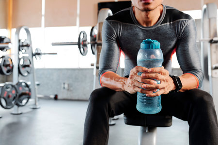 Fit sweaty young man resting on bench with bottle of fresh water after training