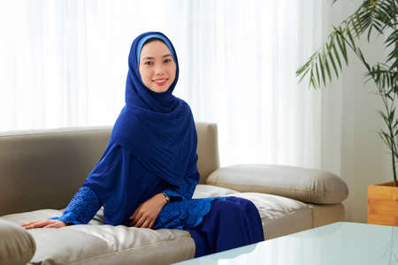 Pretty young Muslim woman in blue press and hijab resting on sofa at homeの写真素材
