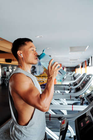 Serious young Vietnamese man standing on treadmill and drinking fresh water before trainingの写真素材