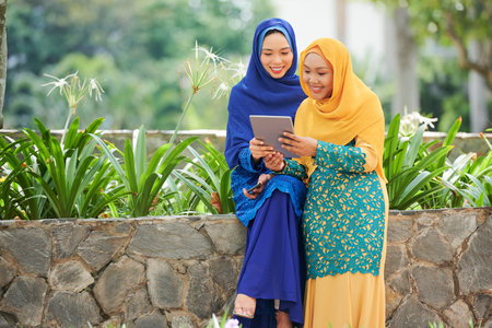 Vietnamese female friends on hijabs standing in park and watching something on digital tabletの写真素材