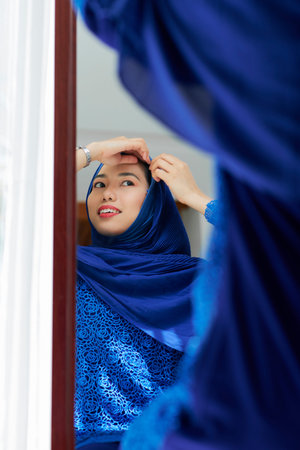 Pretty smiling young Asian woman putting on blue hijab in front of mirrorの写真素材