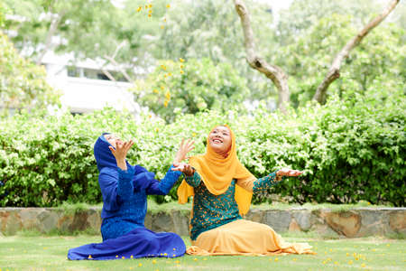 Beautiful positive young Vietnamese woman in traditional muslim dresses sitting on green grass and playing with petalsの写真素材