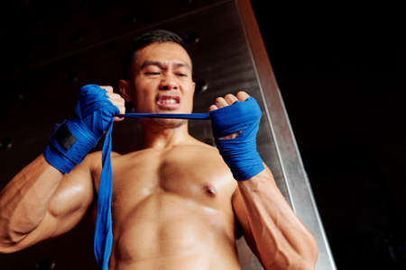 Asian shirtless boxer preparing for fight and wrapping his fists wth angry expression in gymの写真素材