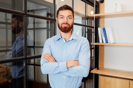 Portrait of young bearded businessman standing in blue shirt with arms crossed and looking at camera...