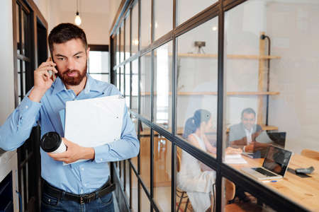 Serious young businessman holding folders and cup of coffee and talking on mobile phone while walking along the office corridor to the meetingの写真素材