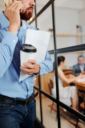 Young bearded male executive talking on mobile phone while carrying folder with documents and coffee drink to the business meeting with his partnersの写真素材