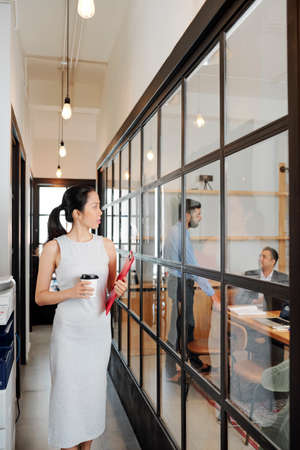 Young Asian office worker holding folder with documents and coffee drink walking along the office corridor and looking at business people working behind the glass wallの写真素材