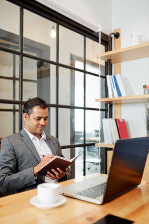 Indian male executive working at his workplace with laptop computer and writing a work plan in notepad at officeの写真素材