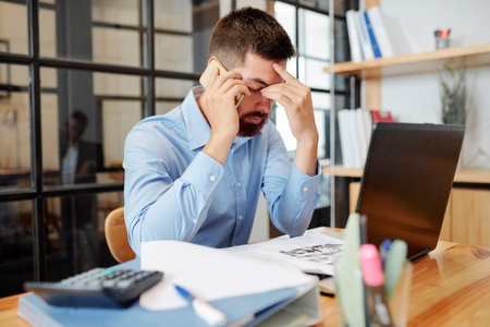 Young bearded businessman sitting at the table in front of laptop talking on mobile phone and look...