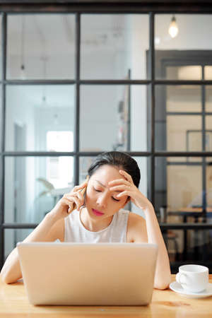 Young office worker sitting at her workplace using laptop computer and talking on mobile phone at officeの写真素材