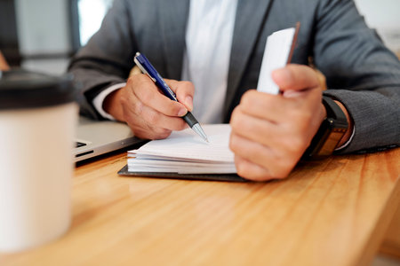 Close-up of businessman sitting at his workplace and making notes with pen in notepad while working...