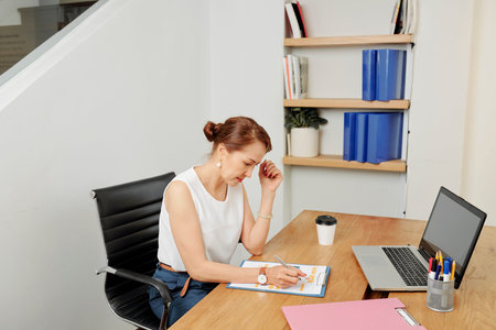 Young businesswoman sitting on chair at the table in front of laptop computer and making notes in...
