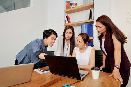 Asian businesswoman pointing at laptop computer and talking about online presentation while her partners listening to her at the table at officeの写真素材