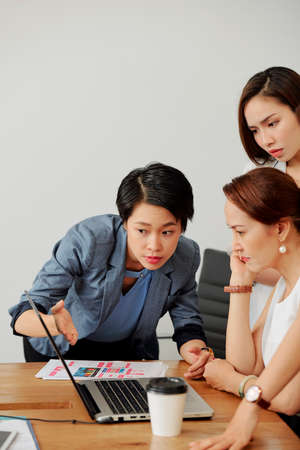 Asian young businesswoman pointing at laptop and showing online project to her female leader while she sitting at the table and looking at it closelyの写真素材