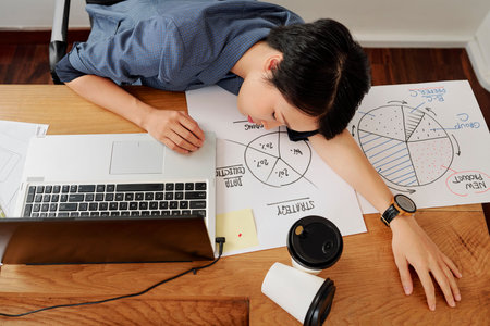 Asian young businesswoman sleeping on her worklace among laptop computer business charts and cups...