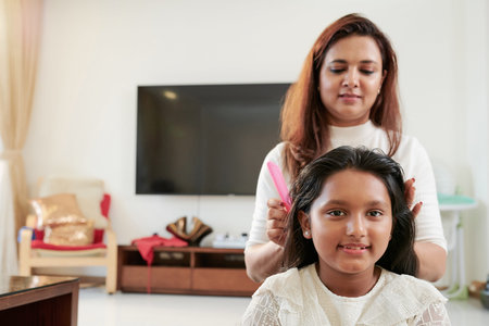 Young girl sitting and looking at camera while mother combing her hair and doing hairstyle in the...