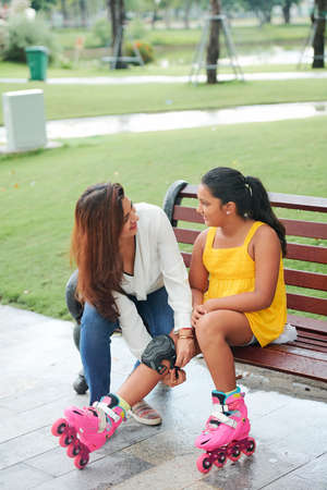 Mother wearing protection on her daughter before skating while they sitting on the bench in the parkの写真素材