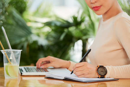 Close-up of young woman making notes in notepad while working on laptop at the table with glass of lemonade in cafeの写真素材