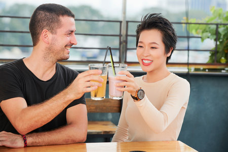 Happy young couple sitting at the table and toasting with glasses of fresh juice during their...