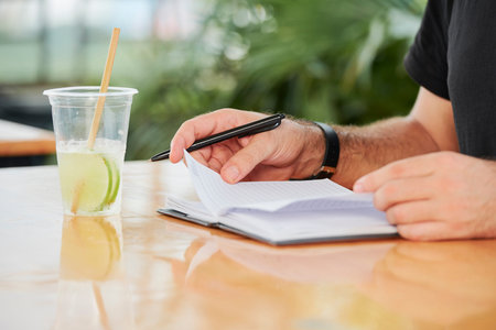 Close-up of young man sitting at the table with fresh cocktail opening his notepad and writing some...