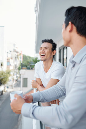 Happy young man laughing at joke of his coworker when they are drinking coffee and chatting at office balconyの写真素材
