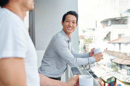 Cheerful young Vietnamese businessman with mug of coffee listening to joke of his colleague