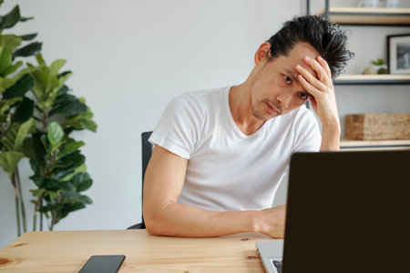 Portrait of bored or stressed Asian man working on laptop at office table