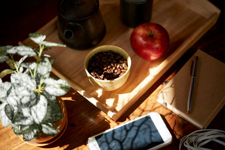 Bowl of coffee beans and fresh apple on tray next to planner and smartphones