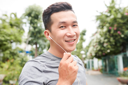 Portrait of cheerful Asian sportsman with earphones in his ears trangin outdoors