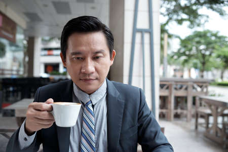 Pensive serious mature Asian businessman with cup of coffee sitting at cafe and reading articles on laptopの写真素材