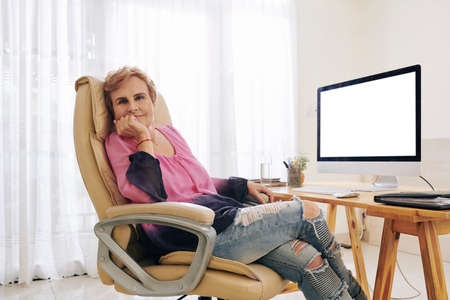 Portrait of smiling tired senior female entrepreneur in casual clothes resting in her office chair and looking at cameraの写真素材