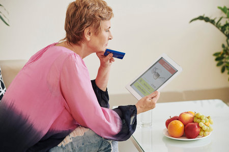 Aged woman with credit card sitting on sofa at home and choosing sunglasses in online store