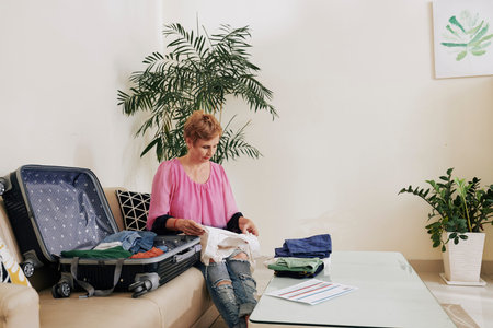 Senior woman sitting at table with plane ticket and stack of clothes and packing suitcase for summer...