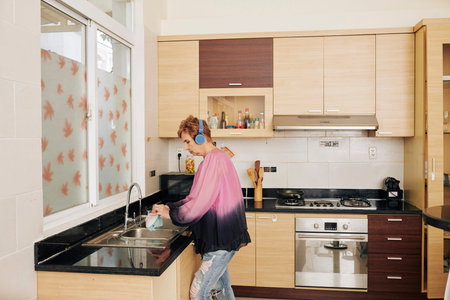 Senior woman in headphones listening to music when washing dishes in her kitchen