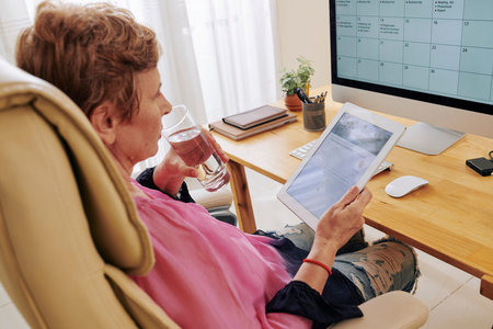 Aged business lady sitting at table, drinking glass of water and reading article on digital tablet
