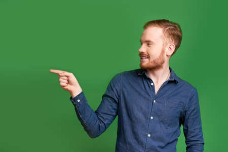 Cheerful young Caucasian man pointing at something with index finger, isolated on greenの写真素材