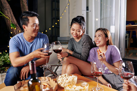 Joyful young Asian people sitting in backyard and enjoying wine with delicious pizzaa