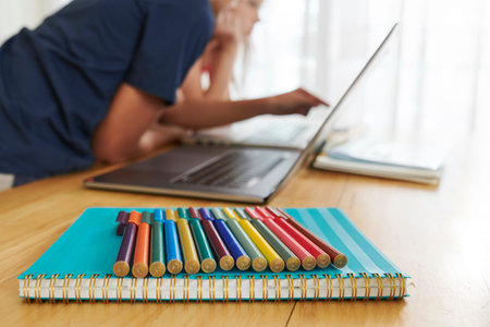 Colorful markers and textbook on desk in school class, children working on laptop in backgroundの写真素材