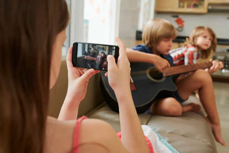 Girl using smartphone to make video of preteen boy singing on sofa and playing guitar at homeの写真素材