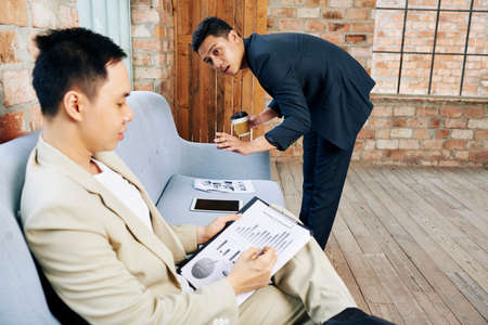 Serious entrepreneur checking report on sofa and talking to coworker who is analyzing financial chartの写真素材