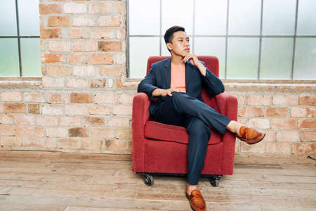 Young confident Asian entrepreneur in stylish suit sitting in velvet armchair and pondering over idea for new businessの写真素材